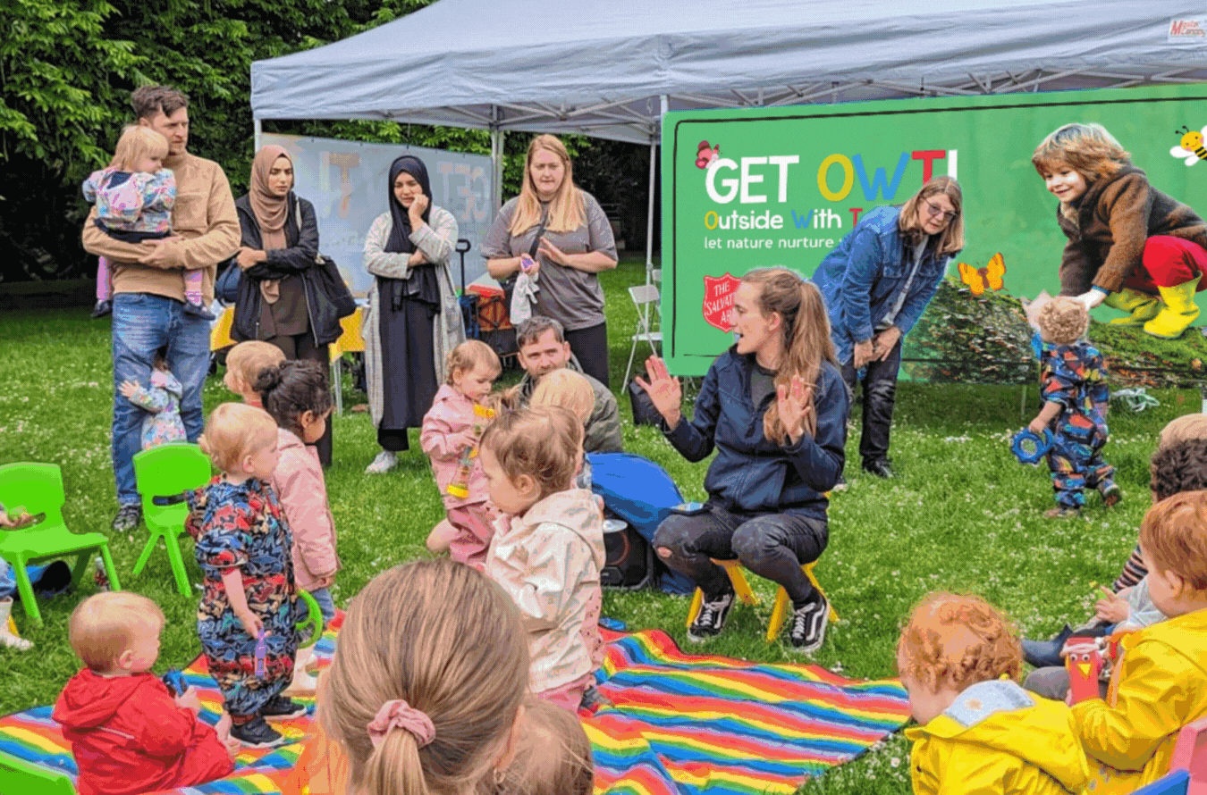 a looping GIF of an outdoor vinyl banner featuring the words 'Get OWT! outside with toddlers' and a boy playing on a log
