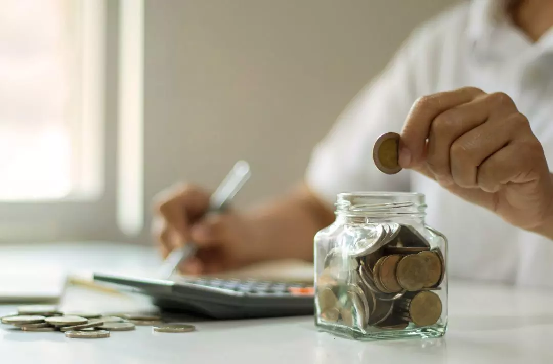 A photo of someone putting coins into a glass jar and making notes. A calculator is on the table.