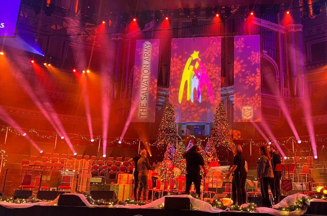 A photo of the stage of the Royal Albert Hall with Christmas trees and banners with The Salvation Army red shield. The production crew is having a planning meeting.