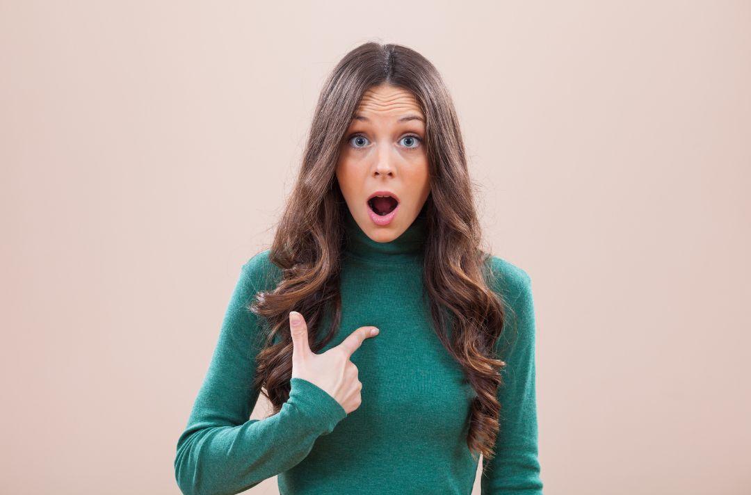 A photo of a woman pointing to herself with her mouth open, as if she is saying 'what, me?'