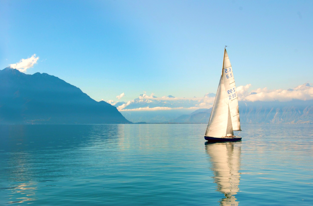 Photo shows a sailing boat on calm waters with hazy mountains in the distance.