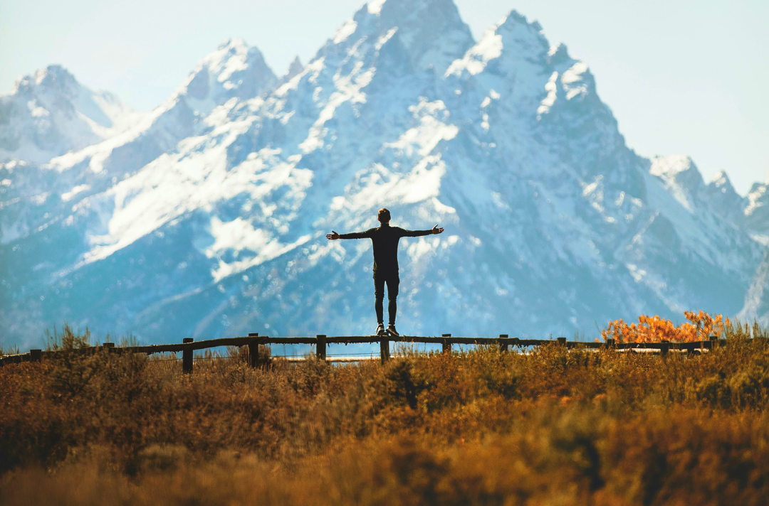 Photo shows someone silhouetted with arms outstretched above a grassy field and in front of a mountain.