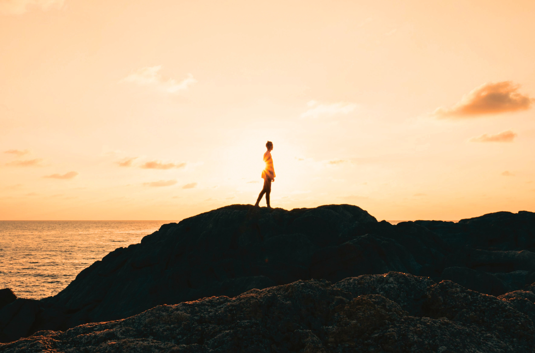 Photo shows someone standing on a rock near the sea silhouetted by the sun.