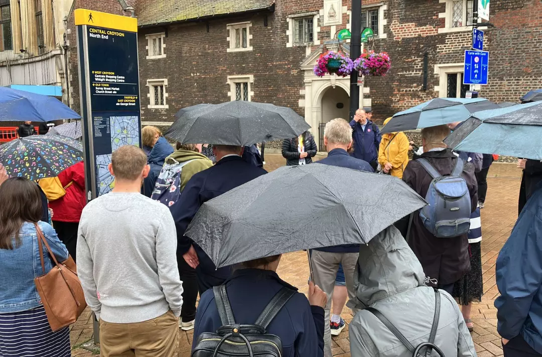 A photo of a large group gathering outside an old building in Croydon, praying in the rain. People are standing under umbrellas.