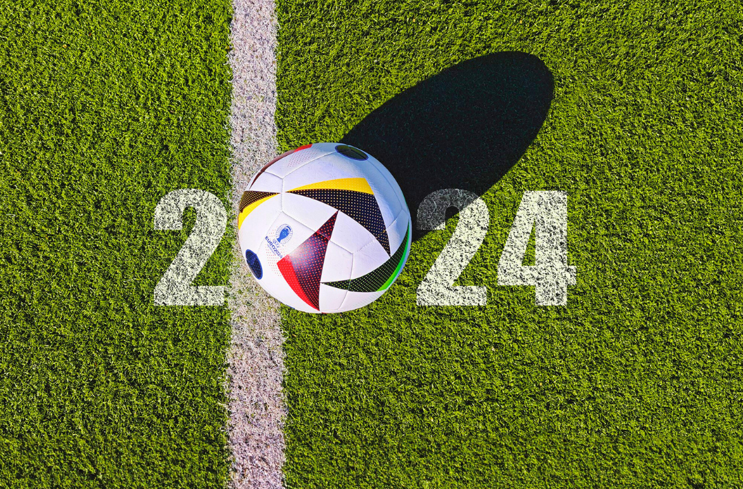 A photo shows a football on a pitch. Below the football, 2024 is written on the grass.