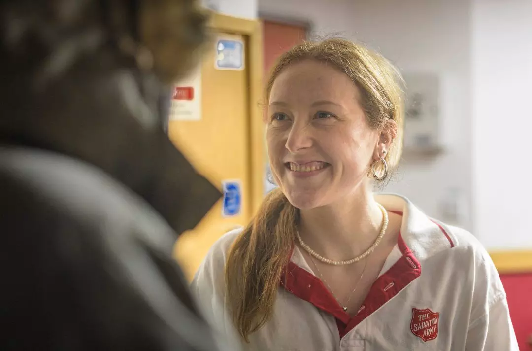 A photo of a lady wearing a Salvation Army polo shirt smiling at someone as she lets them into a Salvation Army building out of the cold