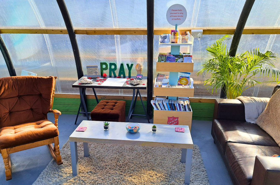 A photo shows the inside of a polytunnel. Two comfy chairs sit beside a table of prayer resources, suggesting a cosy, yet airy, atmosphere.