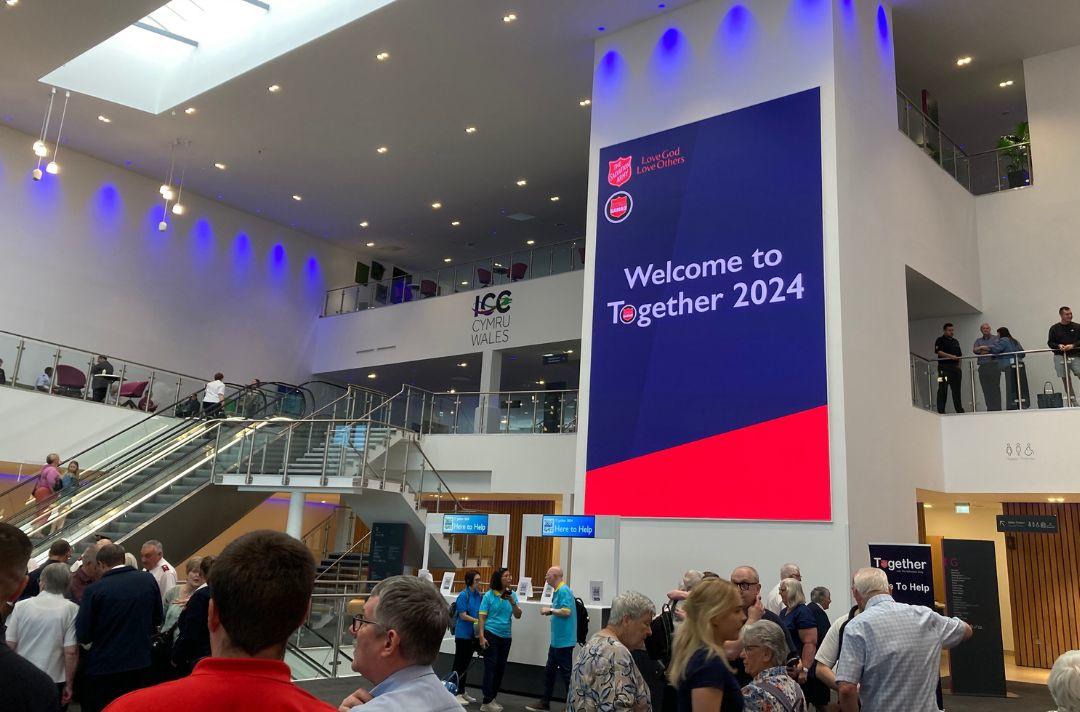A photo of people in the atrium of ICC Wales with a large digital screen that reads Welcome to Together 2024