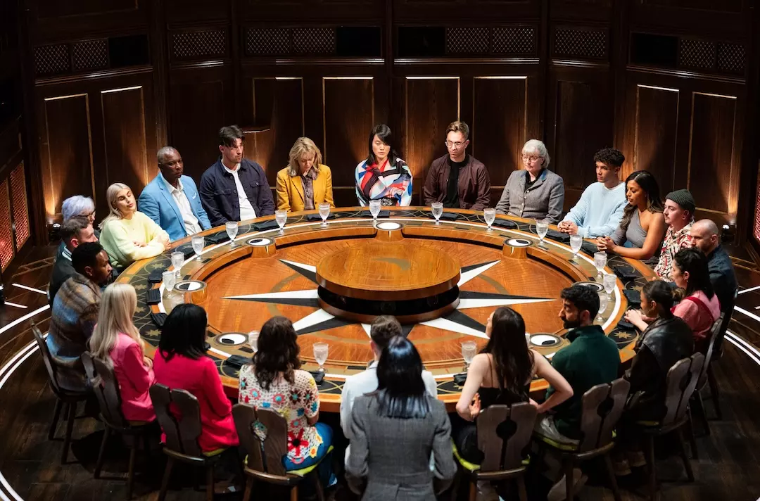 Claudia Winkleman stands by a table around which all the contestants are seated for the 2025 series of The Traitors
